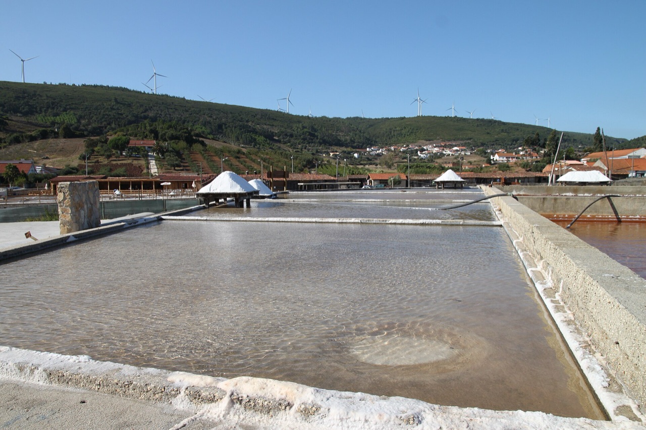 Sunlit salt pans with white salt cones and wind turbines on the green hills behind