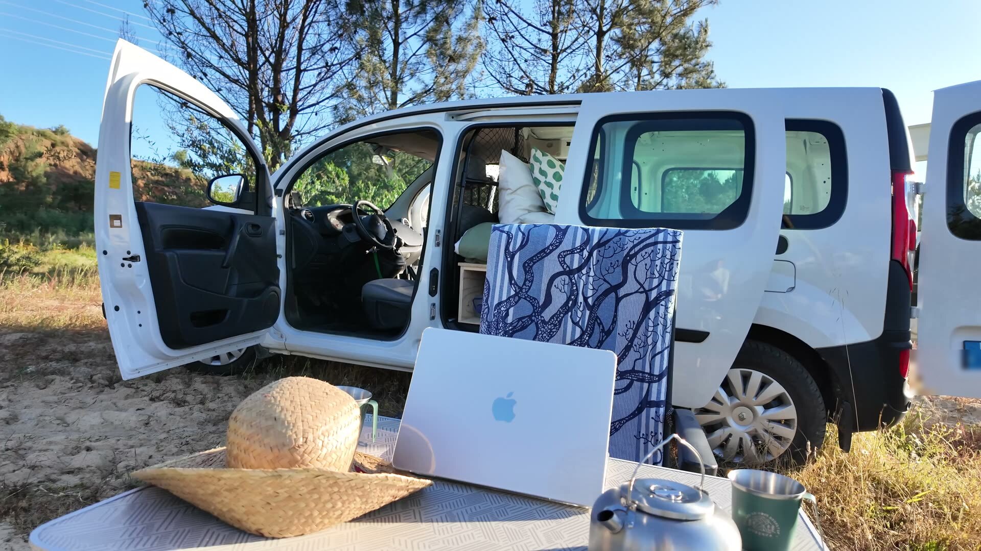 Open van with a camp table holding a MacBook, kettle and straw hat