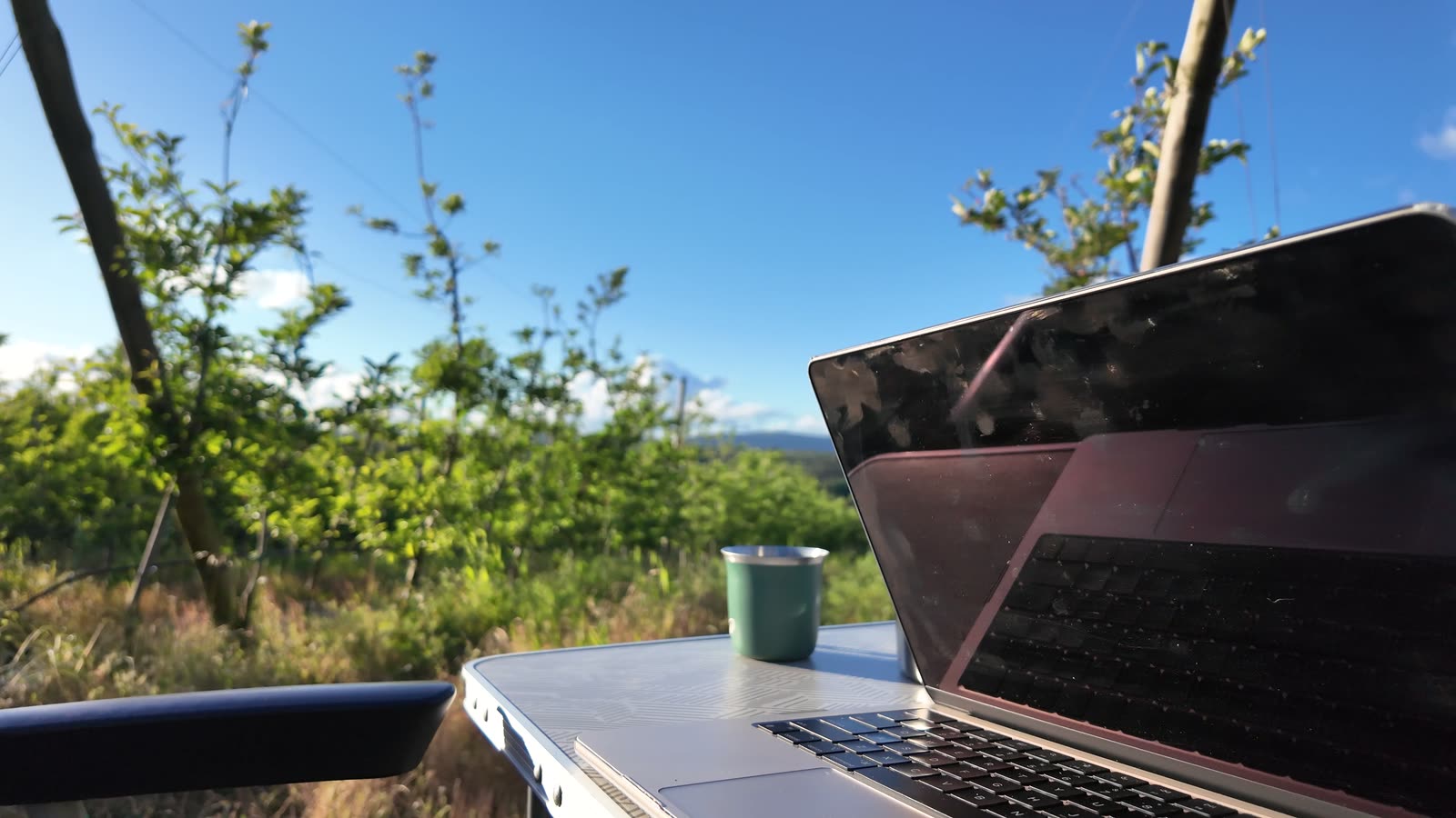 MacBook and enamel mug on the camp table with the green valley behind