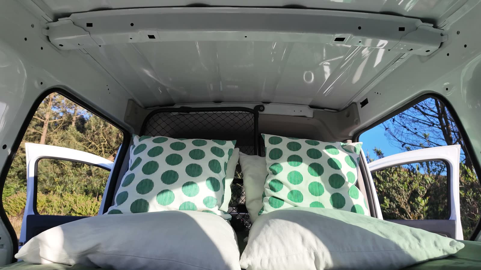 Made bed with green polka-dot pillows and windows opening onto trees and sky